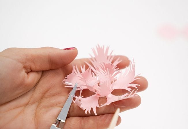 Manos recortando y dando forma a una flor de papel rosa con tijeras y herramienta de modelado, proceso de Cartoncita