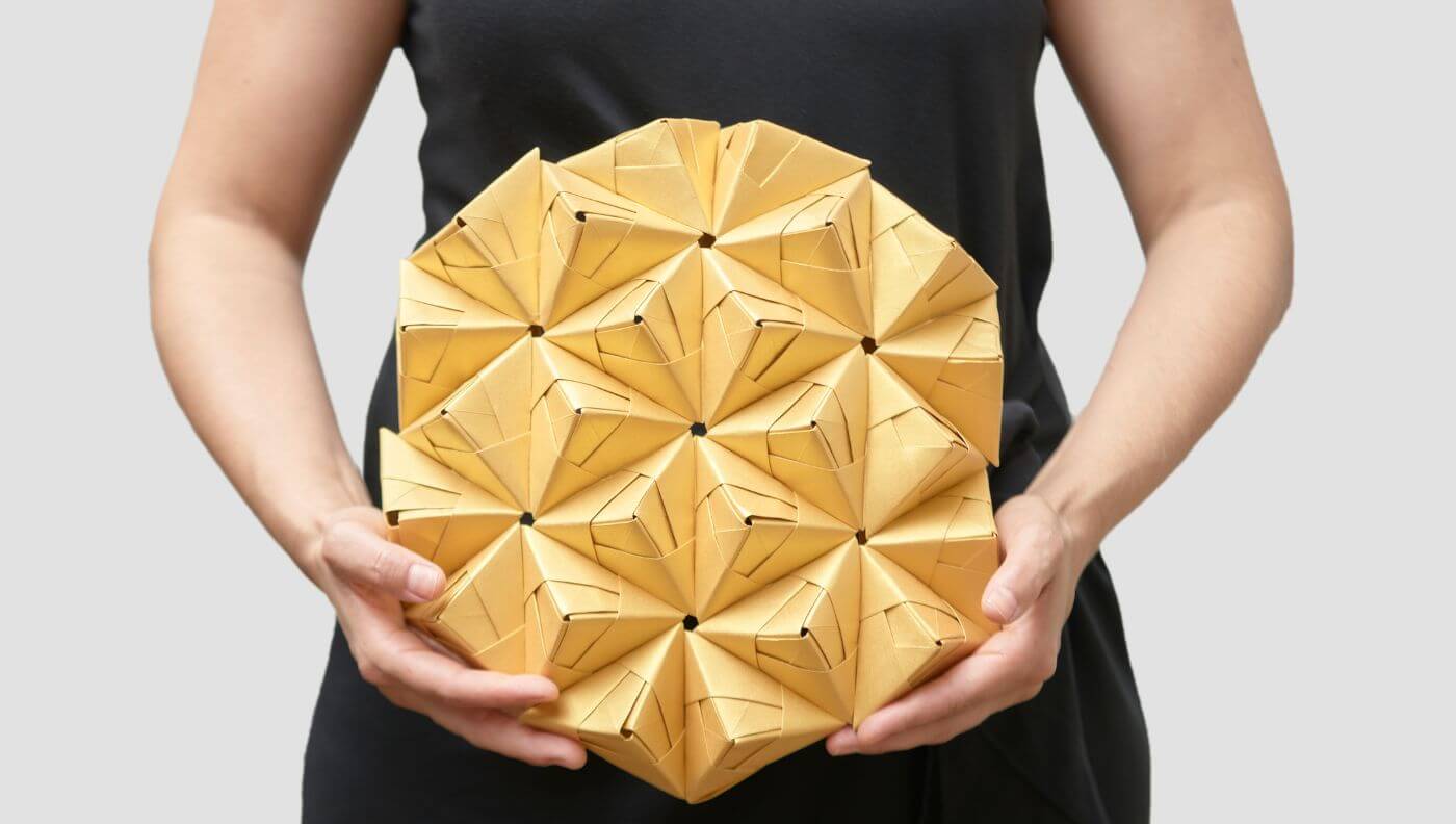 Persona sosteniendo una estructura de papel dorado plegado con pliegues verticales repetidos, diseño de Cartoncita