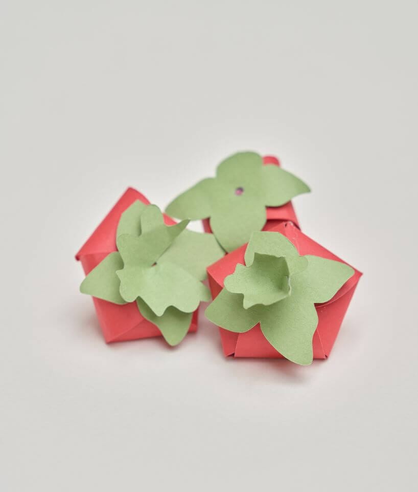 Varias frutas de papel rojas con formas verdes sostenidas entre las manos sobre fondo oscuro, diseño de Cartoncita