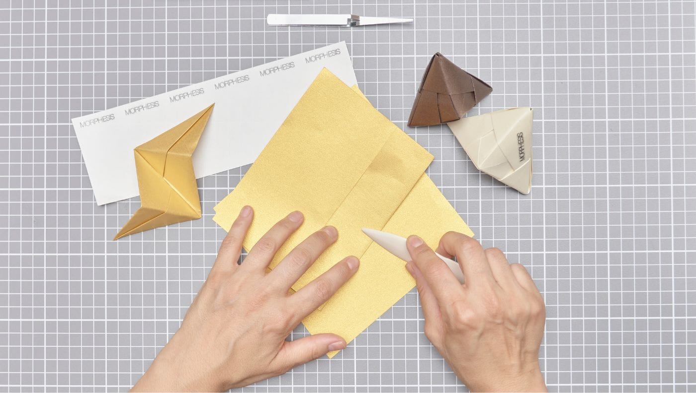 Vista superior de papel dorado siendo marcado y plegado con herramientas sobre una base cuadriculada, trabajo de Cartoncita