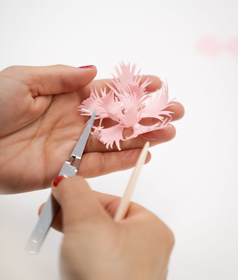 Manos recortando y dando forma a una flor de papel rosa con tijeras y herramienta de modelado, proceso de Cartoncita