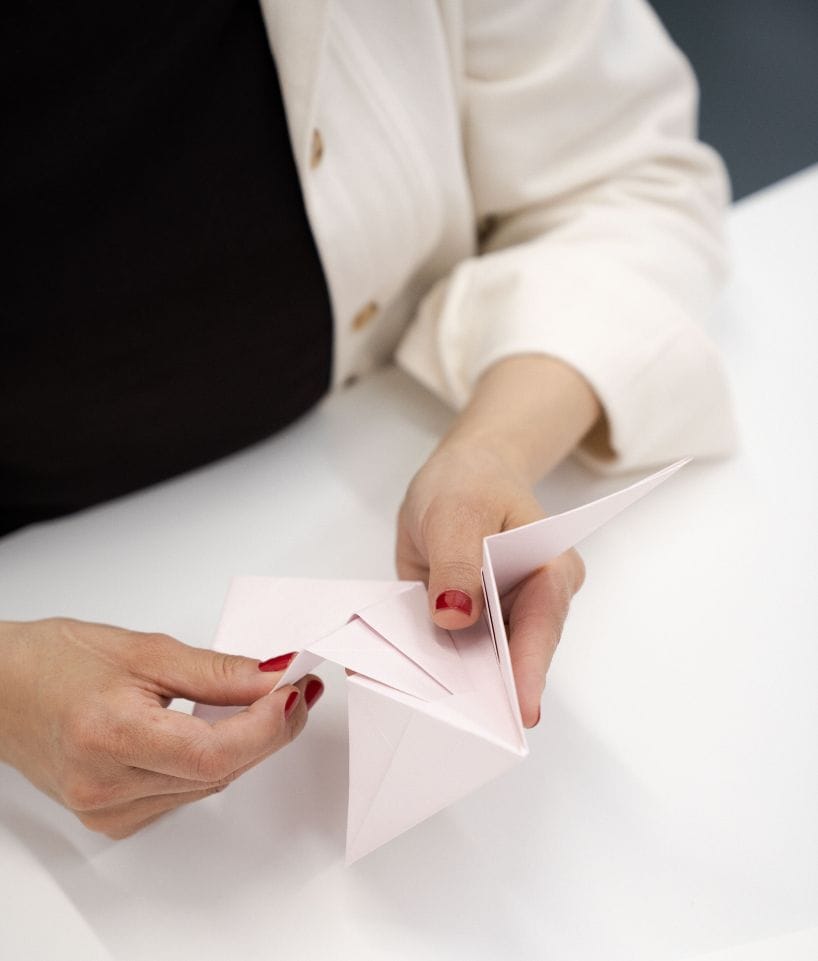 Detalle de manos realizando pliegues en papel blanco para crear una forma geométrica, proceso de Cartoncita