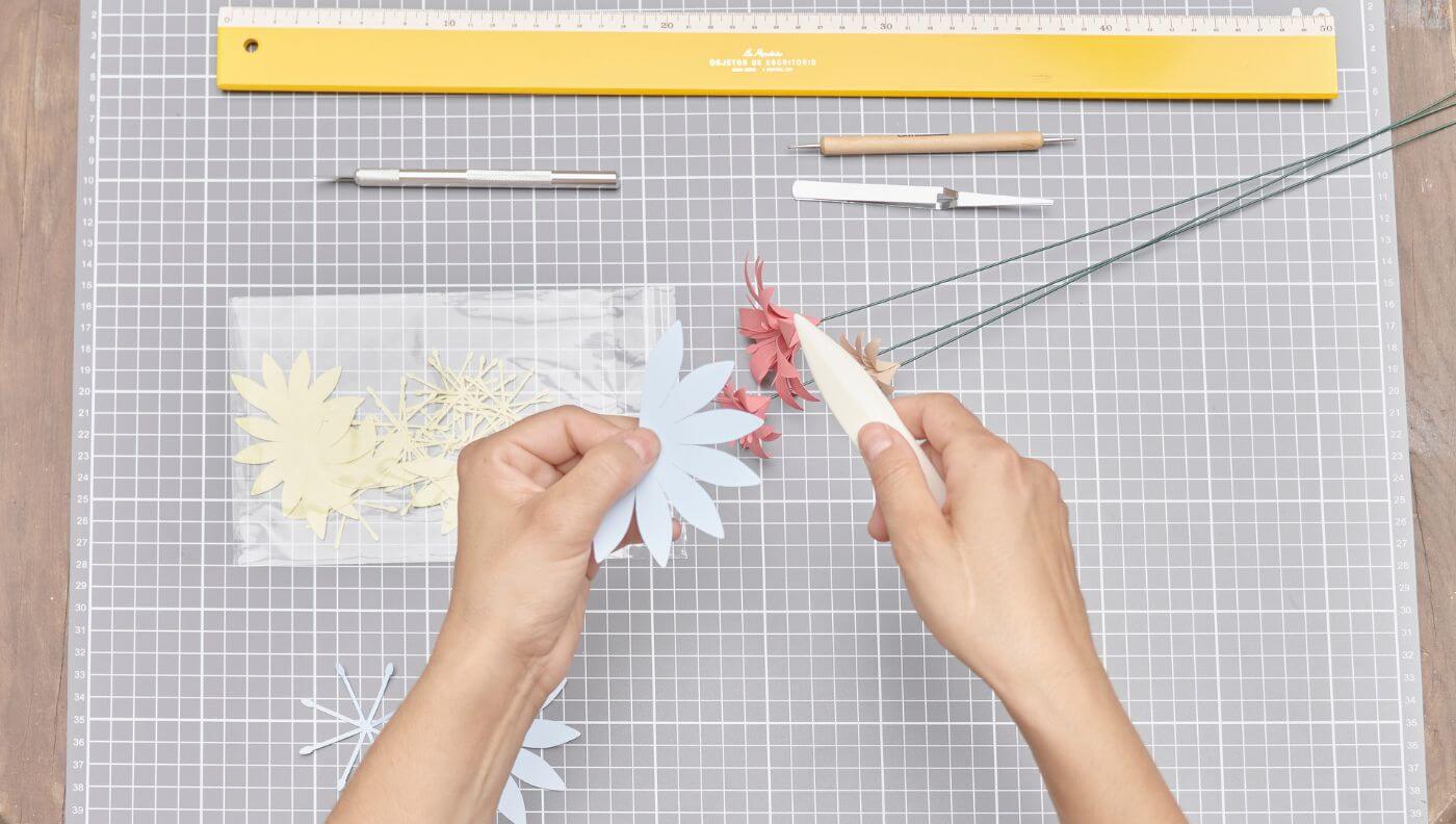 Vista superior del proceso de creación de flores de papel sobre una mesa de trabajo con herramientas, Cartoncita